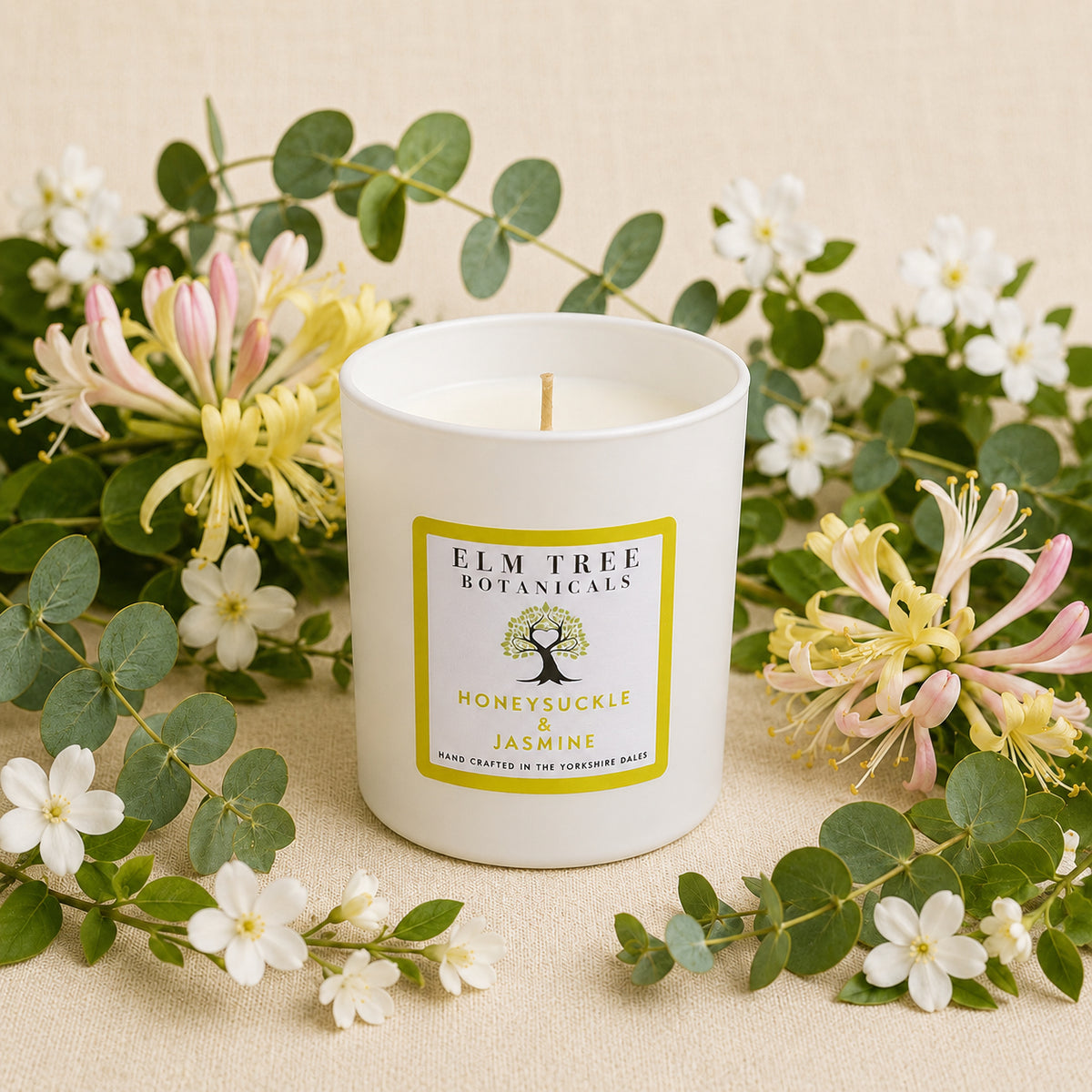 White candle with a label surrounded by flowers and greenery on a beige background