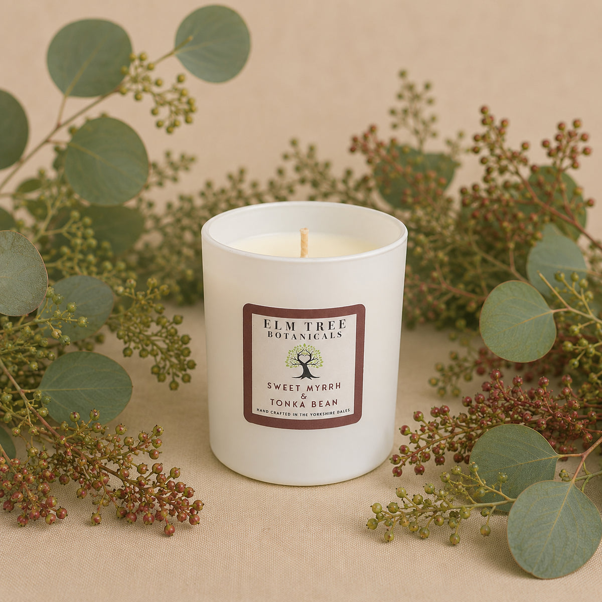 White candle with 'Elm Tree Botanicals' label surrounded by greenery on a beige background