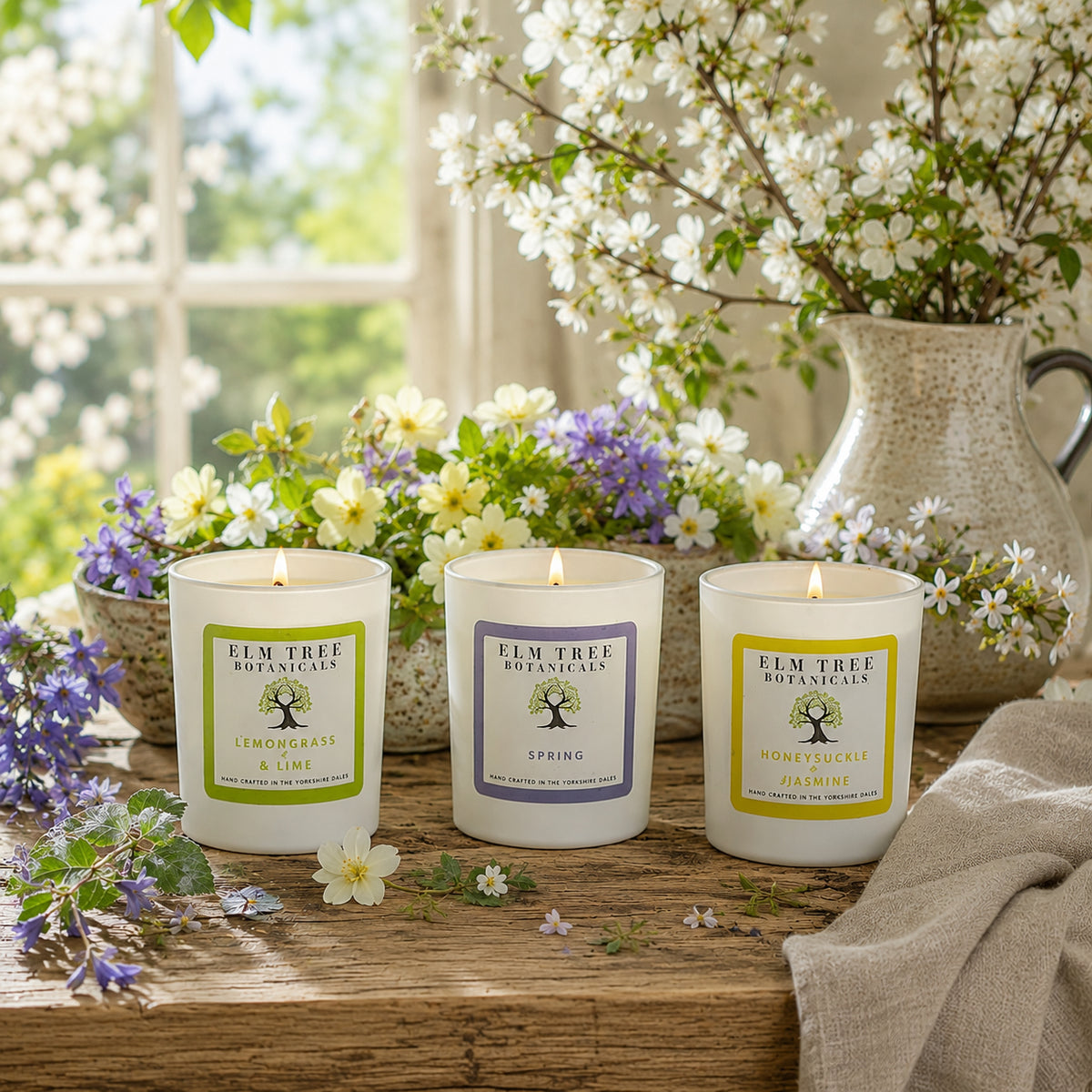 Three lit white candles in glass jars labeled Elm Tree Botanicals are arranged on a wooden table with flowers and a vase in the background.