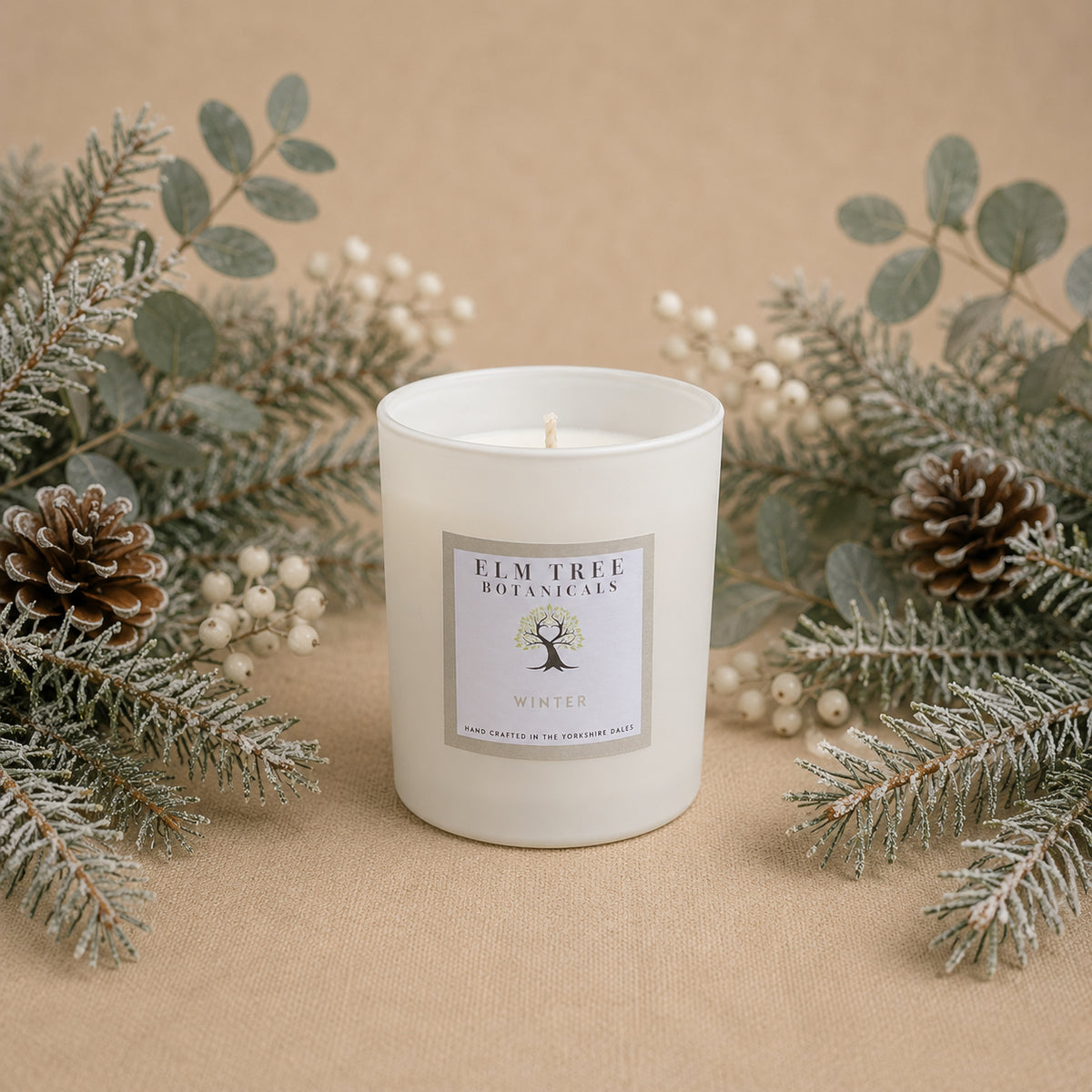 White candle with a label surrounded by pine cones and greenery on a beige background